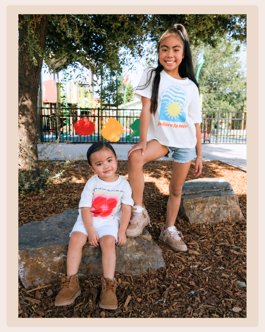Two children wearing casual neutral-colored shoes while posing outdoors near a playground, one standing on a rock and one sitting beside her.