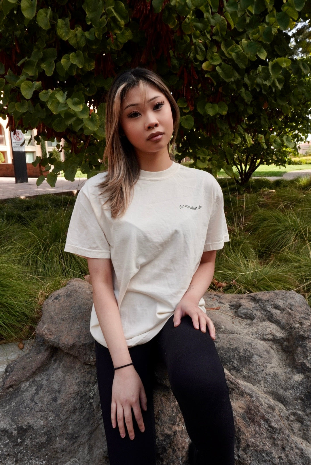 A woman with blonde-highlighted hair sits on a large rock outdoors, wearing a cream-colored t-shirt with a small dark logo on the left chest and black leggings. The background features green foliage and grass.