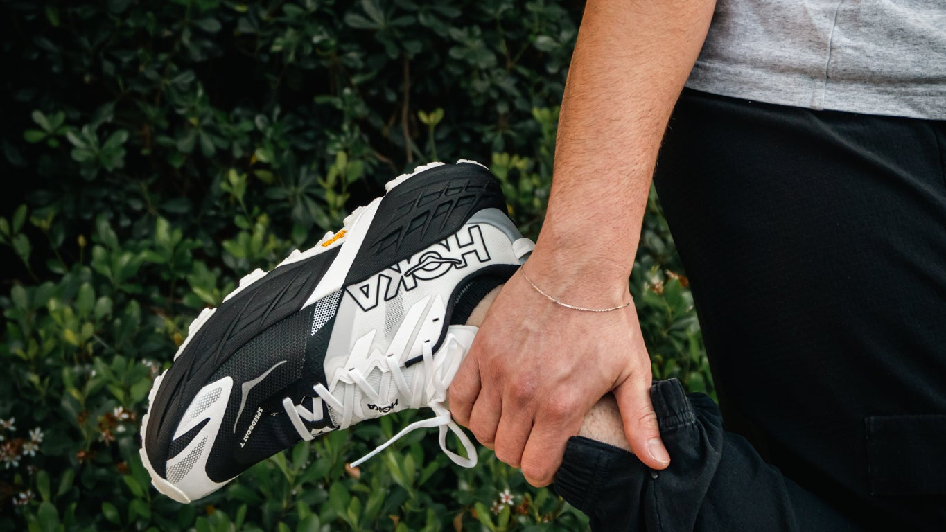 A person outdoors wearing a gray t-shirt and black pants, stretching their leg while holding up a black and white Reebok sneaker.