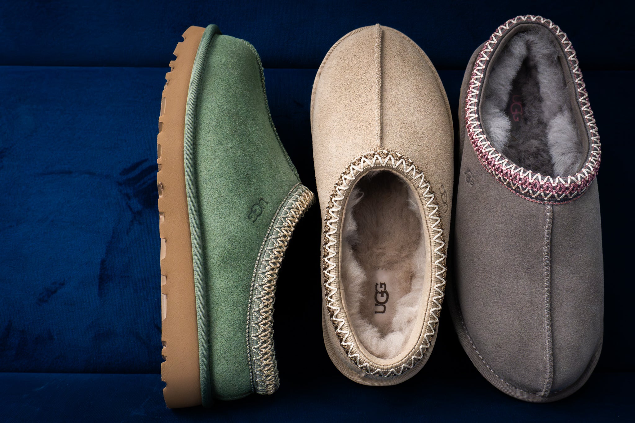 Three UGG suede slippers in green, beige, and gray with soft shearling lining and decorative stitching, displayed against a dark blue background.