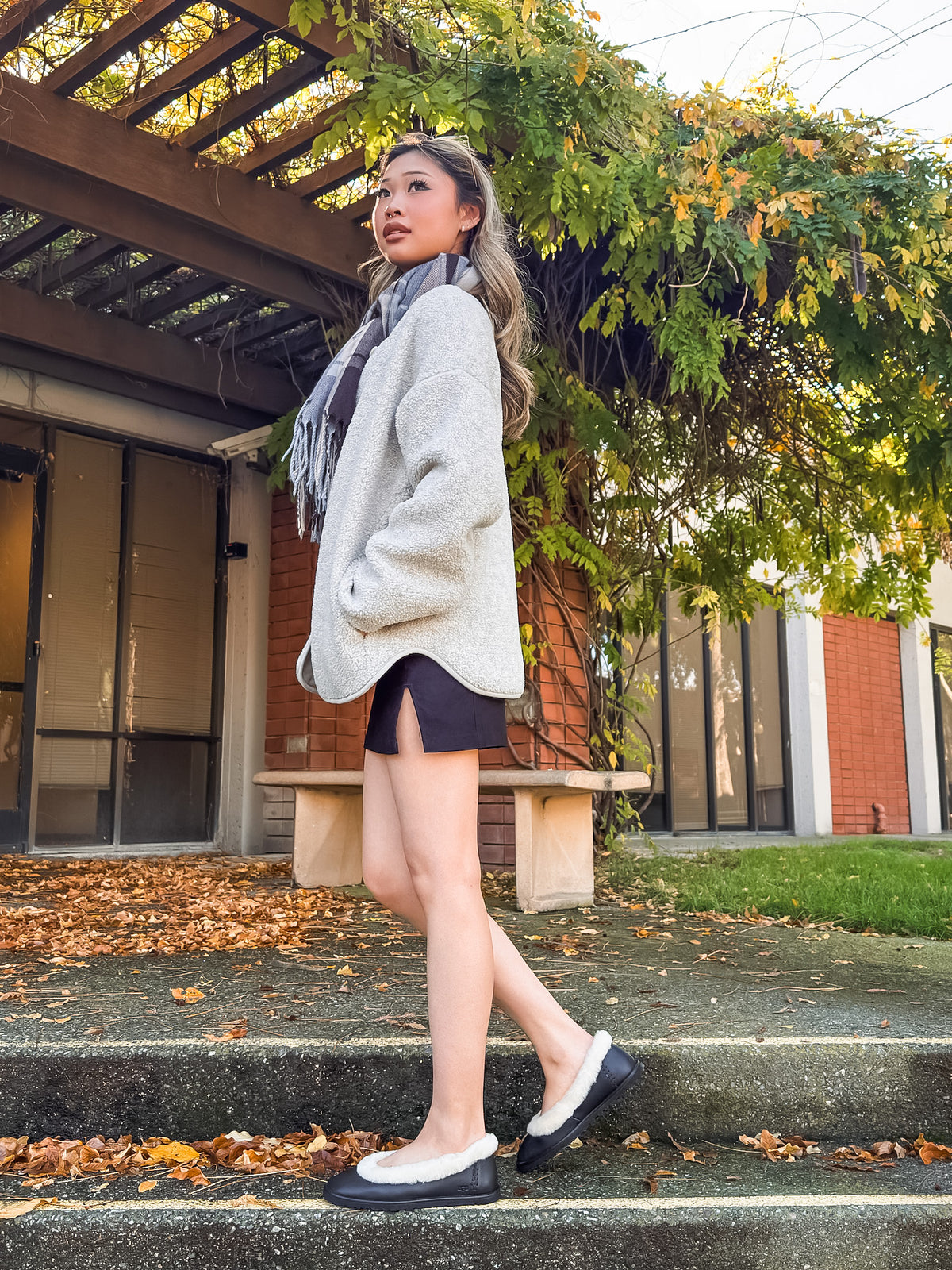 A woman wearing a light gray oversized knit cardigan, black shorts, a gray scarf, and black-and-white loafers walks down concrete steps outdoors in front of a brick building with greenery and fallen leaves.