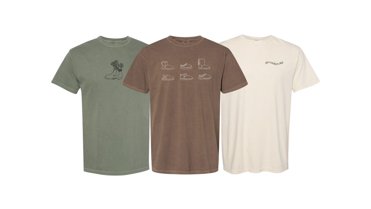 Three short-sleeve crewneck t-shirts displayed side by side on a plain white background: one sage green, one medium brown, and one cream. Each shirt features a minimalist line-art graphic on the chest, including boots and shoes.