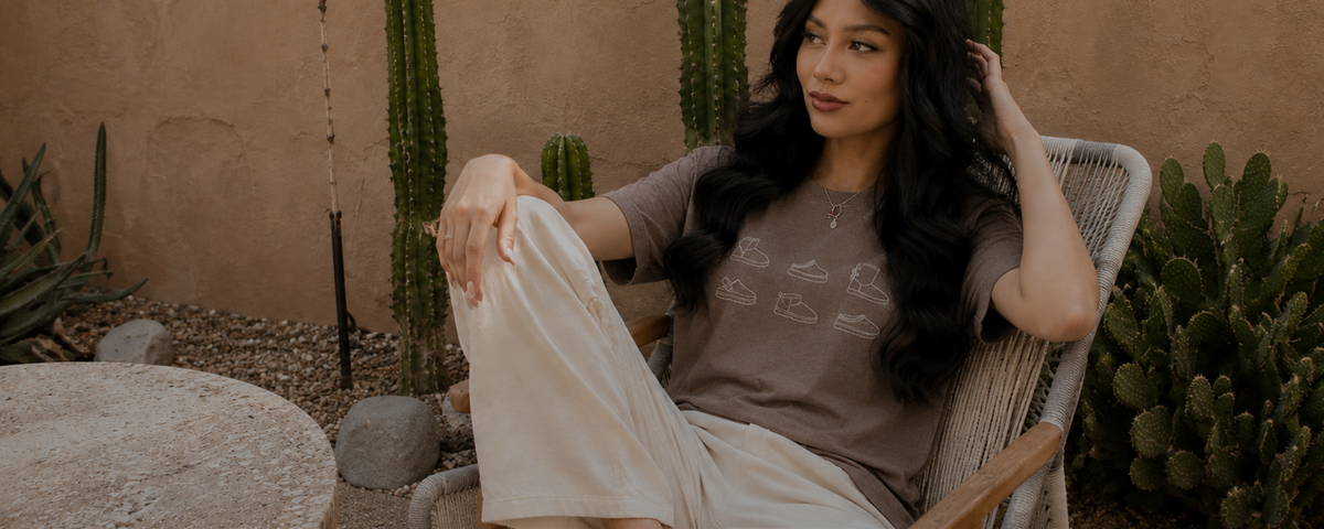 A woman with long dark hair sits in a woven outdoor chair in a desert garden setting, wearing a taupe graphic t-shirt and cream wide-leg pants.